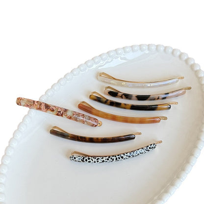 Seven slim French barrettes in assorted acetate colors and patterns arranged on a white dish
